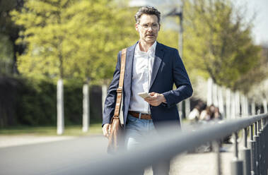 Male entrepreneur with shoulder bag and mobile phone walking by railing - UUF23194