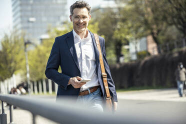Smiling businessman holding mobile phone while standing on sunny day - UUF23195