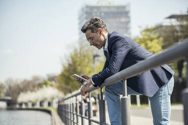 Älterer Geschäftsmann, der ein Mobiltelefon benutzt, während er sich an einem sonnigen Tag an ein Geländer lehnt - UUF23198