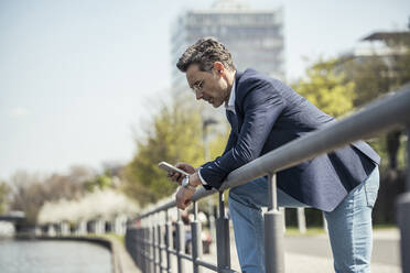 Mature businessman using mobile phone while leaning on railing during sunny day - UUF23198