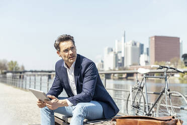 Thoughtful businessman with digital tablet sitting on sunny day - UUF23203