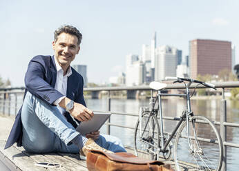 Mature businessman with digital tablet sitting by bicycle during sunny day - UUF23208