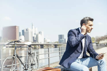 Mature male entrepreneur adjusting in-ear headphones while sitting on sunny day - UUF23210