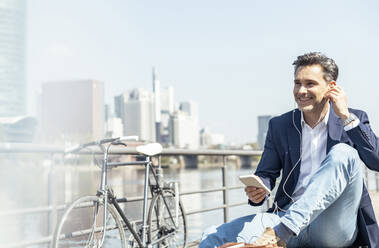 Smiling businessman sitting with mobile phone and in-ear headphones during sunny day - UUF23211