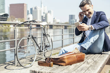 Smiling mature businessman with mobile phone adjusting in-ear headphones while sitting on bench - UUF23212