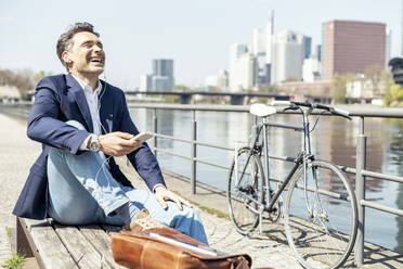 Happy mature businessman sitting by river during sunny - UUF23214