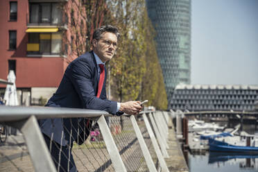 Geschäftsmann mit Mobiltelefon, der sich an ein Geländer lehnt, während eines sonnigen Tages - UUF23219