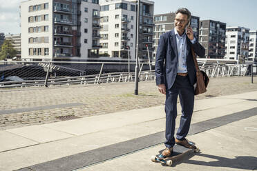 Lächelnder männlicher Unternehmer, der mit seinem Handy telefoniert, während er in der Stadt Skateboard fährt - UUF23234