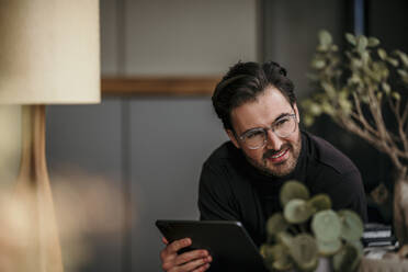 Smiling male professional looking away while holding digital tablet at home - MJRF00554