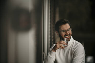 Lächelnder Mann, der ein Glas Wasser in der Hand hält, während er zu Hause wegschaut - MJRF00557