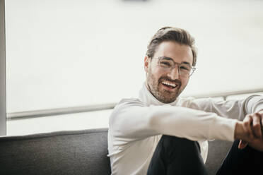 Smiling man sitting in front of bright window at home - MJRF00563