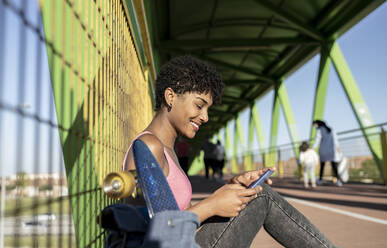 Lockenköpfige Frau benutzt ihr Smartphone, während sie auf einer Brücke am Zaun sitzt - JCCMF02486