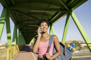 Smiling woman with backpack talking on smart phone at bridge - JCCMF02490