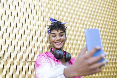 Lächelnde junge Frau, die ein Selfie mit ihrem Smartphone an der Wand macht - JCCMF02529