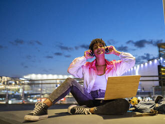 Smiling woman enjoying music on headphones while sitting outdoors at night - JCCMF02562