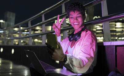 Cheerful woman showing victory sign during video call on smart phone at night - JCCMF02565