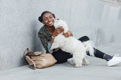 Lächelnde Frau, die mit ihrem Hund spielt, während sie auf dem Gehweg sitzt - JRVF00624
