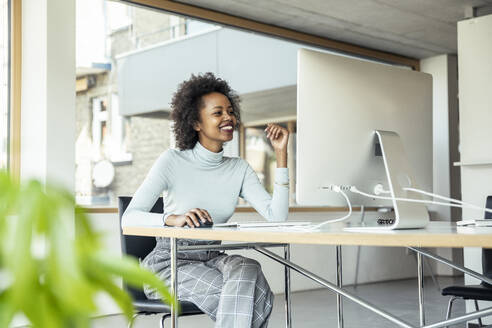 Eine fröhliche Frau, die an ihrem Schreibtisch im Büro mit einem strahlenden Lächeln auf dem Gesicht am Computer arbeitet - UUF23247