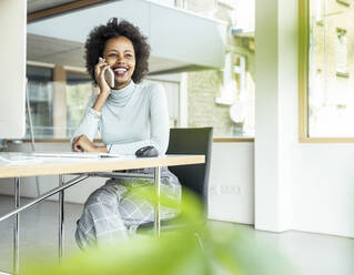 Eine fröhliche Geschäftsfrau, die an ihrem Schreibtisch in einer professionellen Umgebung Multitasking am Telefon betreibt - UUF23260