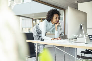 Eine konzentrierte Geschäftsfrau, die an ihrem Computer Multitasking betreibt, während sie sich an ihren Schreibtisch in einer professionellen Büroumgebung lehnt - UUF23263