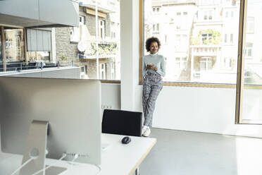 Businesswoman with mobile phone leaning on window at office - UUF23266