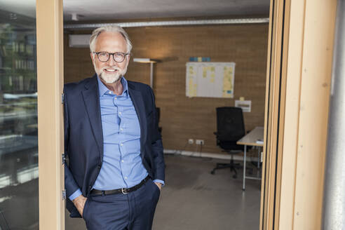 Smiling male professional with hands in pockets leaning on doorframe at office - UUF23297