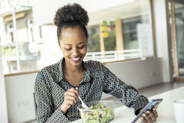 Eine berufstätige Frau, die mit einem Lächeln multitaskingfähig ist und einen gesunden Salat isst, während sie an ihrem Schreibtisch ihr Smartphone benutzt - UUF23300