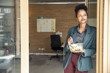 Businesswoman with bowl looking away while leaning on doorframe at office - UUF23301