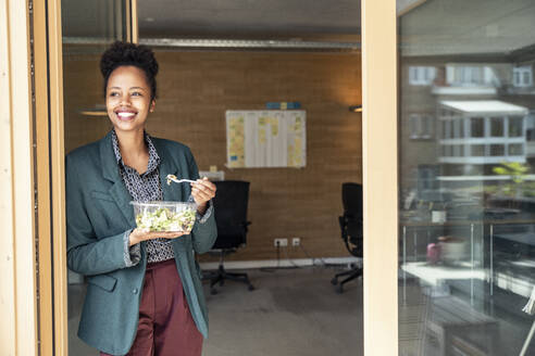 Eine fröhliche Frau genießt einen gesunden Salat, während sie mit einem Lächeln im Gesicht zum Eingang blickt - UUF23302
