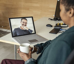 Businesswoman with coffee cup during video conference through laptop at desk - UUF23312