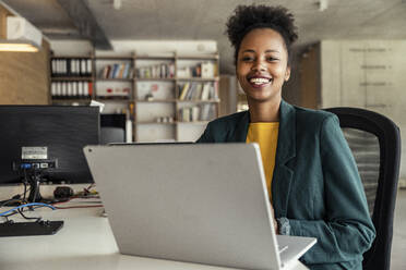 Geschäftsfrau sitzt lächelnd vor einem Laptop im Büro - UUF23317