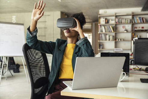 Eine berufstätige Frau benutzt ein Virtual-Reality-Headset und macht Handgesten, während sie an ihrem Schreibtisch im Büro sitzt - UUF23320