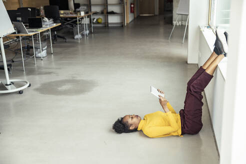 Eine Frau in einem professionellen Umfeld entspannt sich mit hochgelegten Füßen, während sie ein digitales Tablet auf dem Boden benutzt - UUF23327