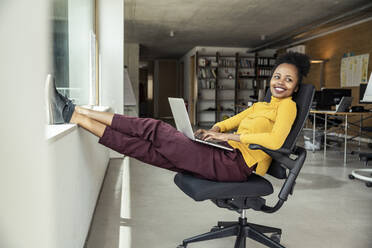 Smiling female professional with laptop looking away while sitting on chair at office - UUF23335