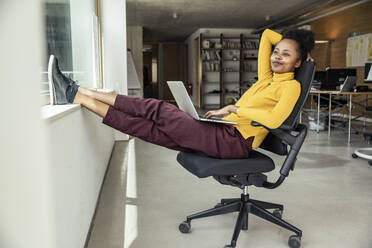 Businesswoman with arm raised looking away while sitting on chair at apartment - UUF23337