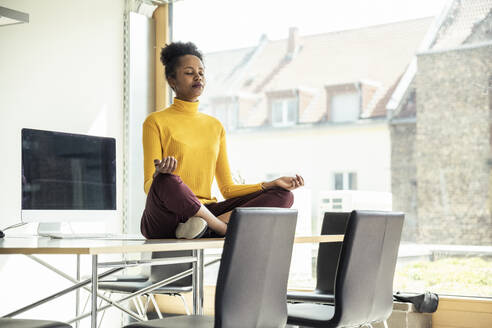 Female professional in lotus position sitting on desk at office - UUF23344