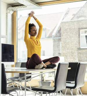 Female professional with arms raised meditating on desk at office - UUF23347