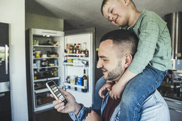 Lächelnder Mann mit Mobiltelefon und Sohn auf der Schulter in der Küche zu Hause - UUF23359