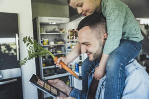 Smiling man using tablet while son sitting on shoulder in kitchen at home - UUF23361