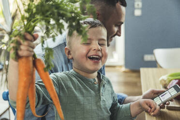 Lächelnder Junge mit geschlossenen Augen, der Karotten hält, während sein Vater zu Hause ein Mobiltelefon benutzt - UUF23376
