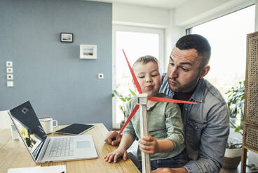 Male freelancer with son blowing wind turbine while playing at home - UUF23378