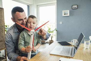 Smiling father sitting with son blowing wind turbine at home - UUF23379