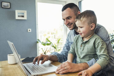 Lächelnder männlicher Freiberufler, der einen Laptop benutzt, während er mit seinem Sohn zu Hause sitzt - UUF23381