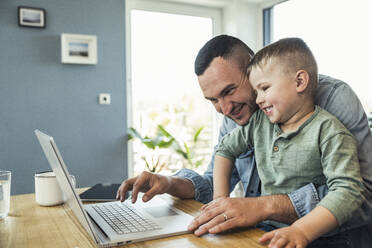 Cheerful businessman using laptop whiles sitting with son at home - UUF23382