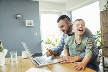 Cheerful boy sitting with father using laptop while working from home - UUF23383