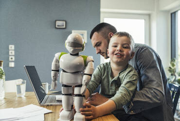 Lächelnder Junge spielt mit einem Roboter, während sein Vater zu Hause am Laptop arbeitet - UUF23384