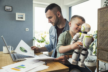 Junge spielt mit einem sitzenden Roboter, während ein Geschäftsmann eine Grafik zu Hause analysiert - UUF23385