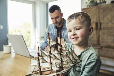 Cute boy playing with toy boat while father working on laptop at home - UUF23386