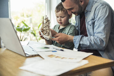 Boy with toy boat sitting with father working from home - UUF23387