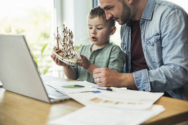 Male freelancer looking at son playing with toy boat at home - UUF23388